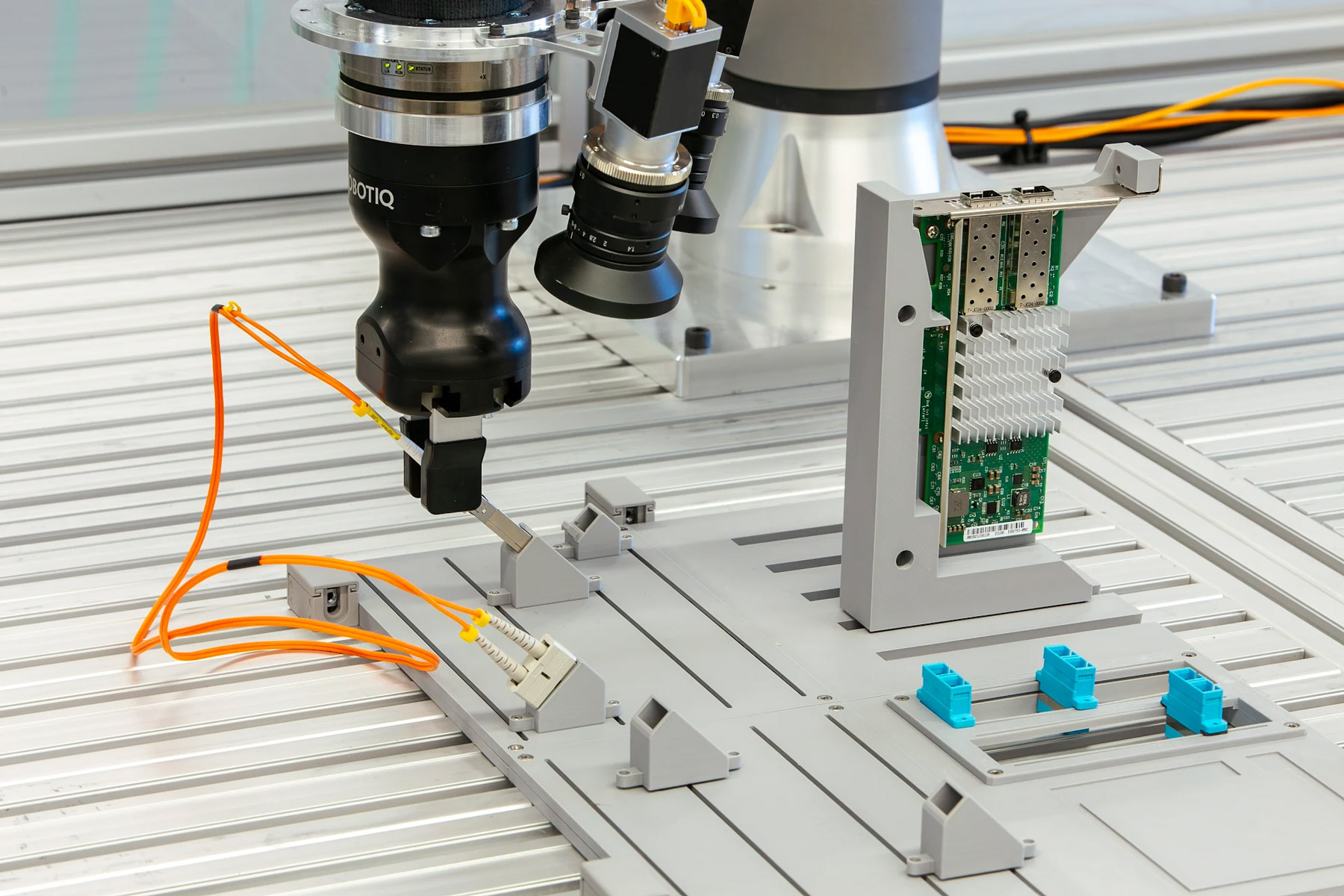Official Intrinsic AI for Industry Challenge workcell showing a robot gripper, orange cables and an electronics board.